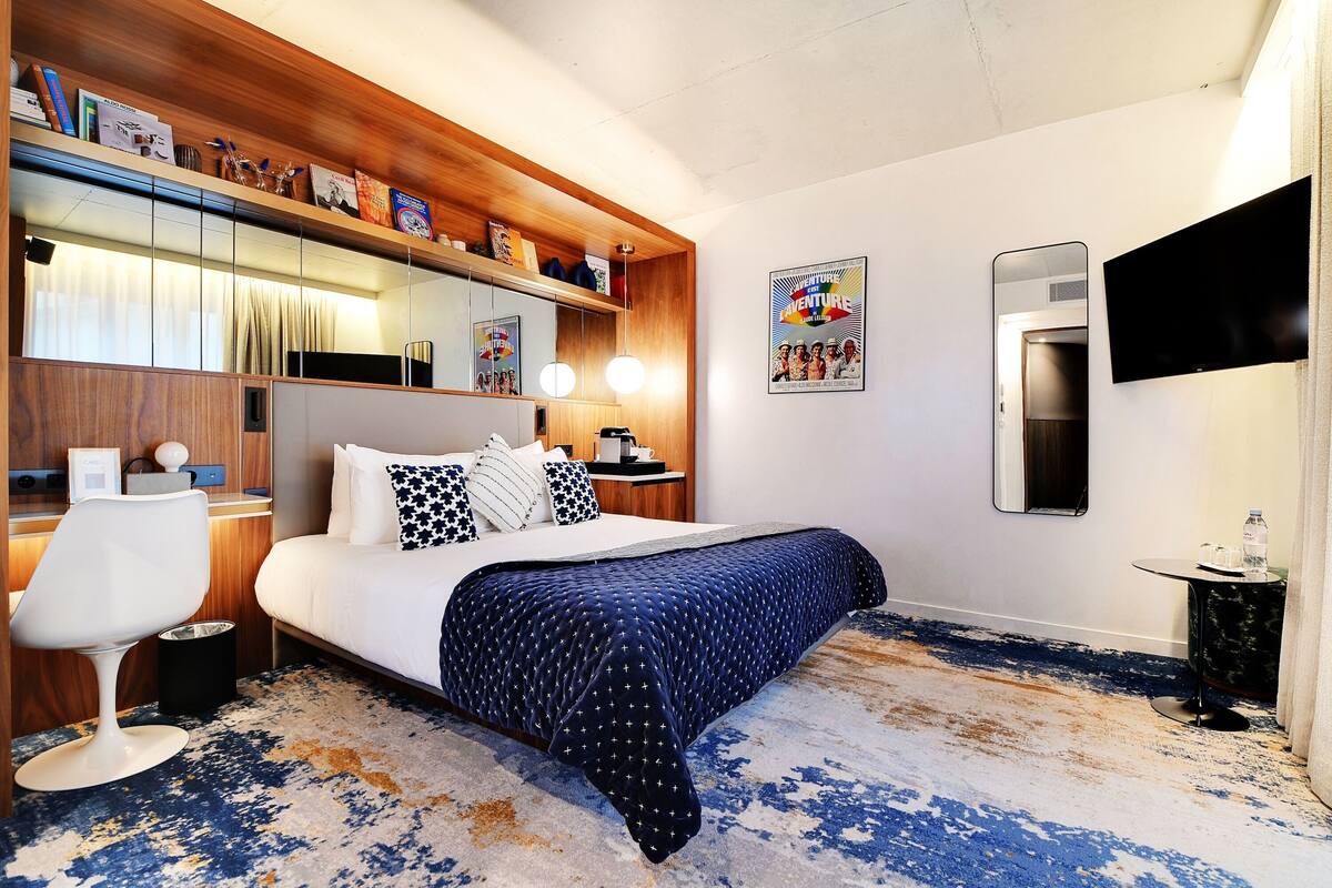 A hotel room with a bed, a desk, a chair, a bookshelf, a mirror, and a TV.