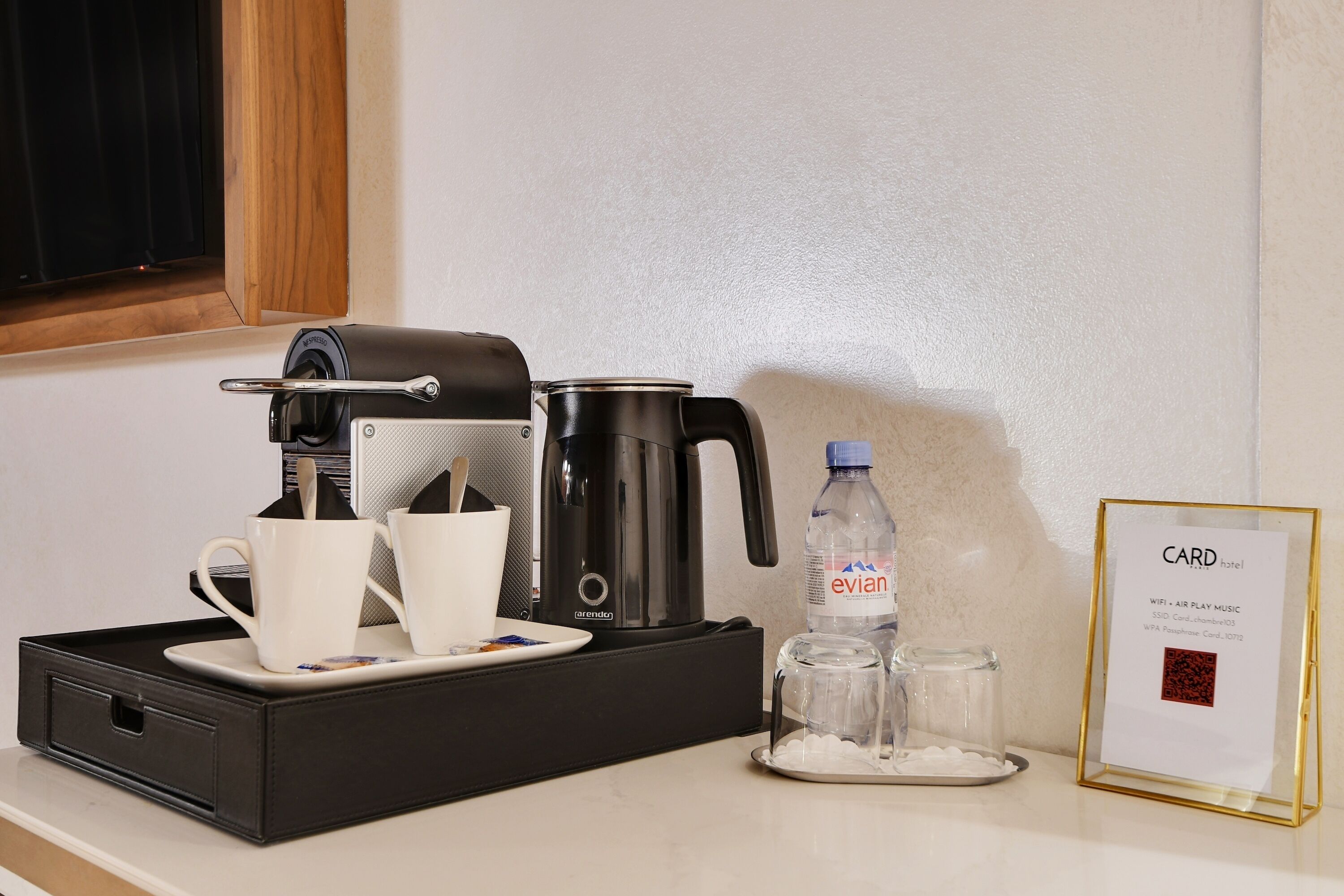 A hotel room with a coffee maker, cups, a water bottle, glasses, and a card detailing amenities like WiFi and airplay music.