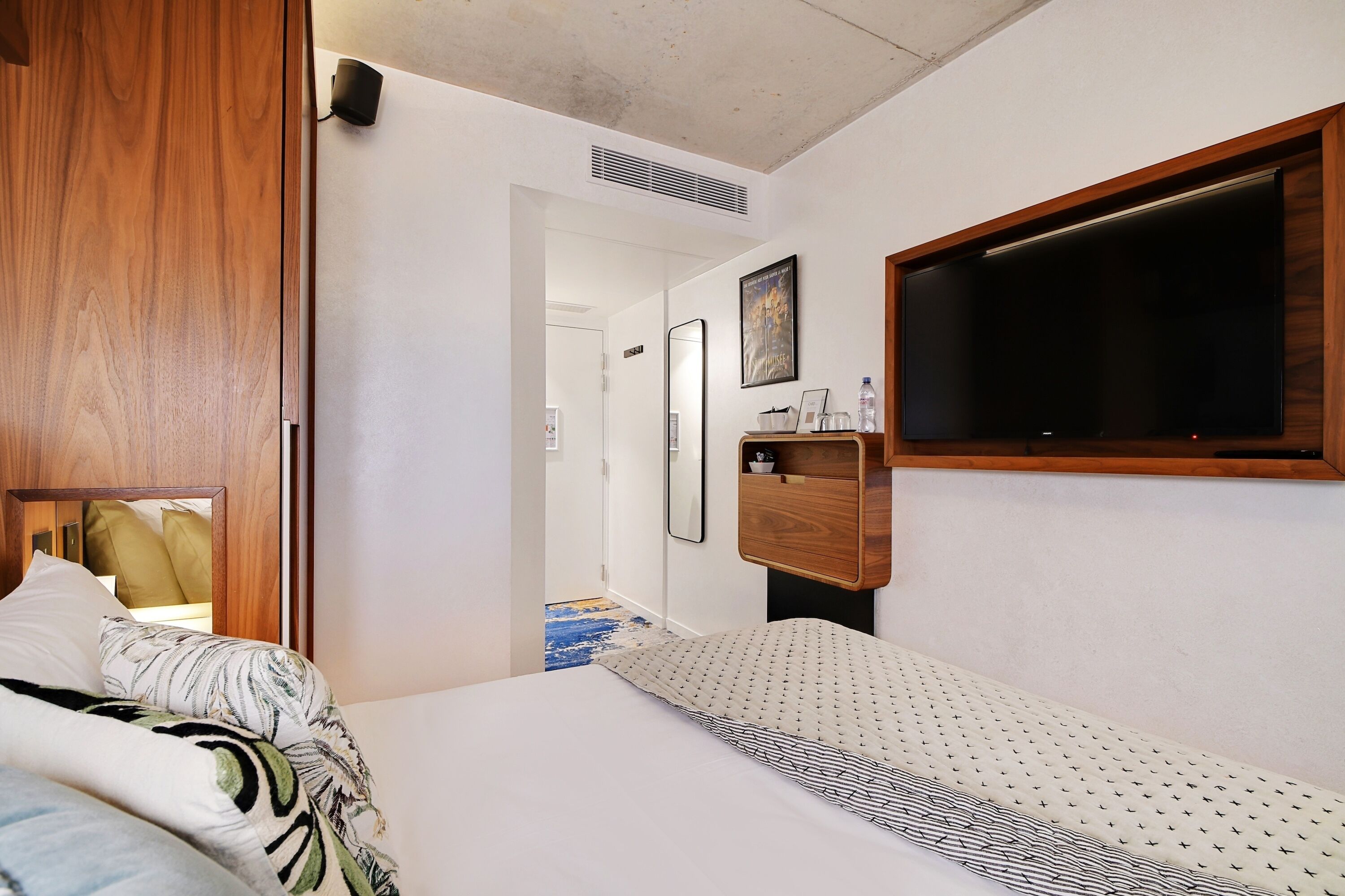 A hotel room with a bed, a wooden wardrobe, a mirror, a shelf with toiletries, and a window.