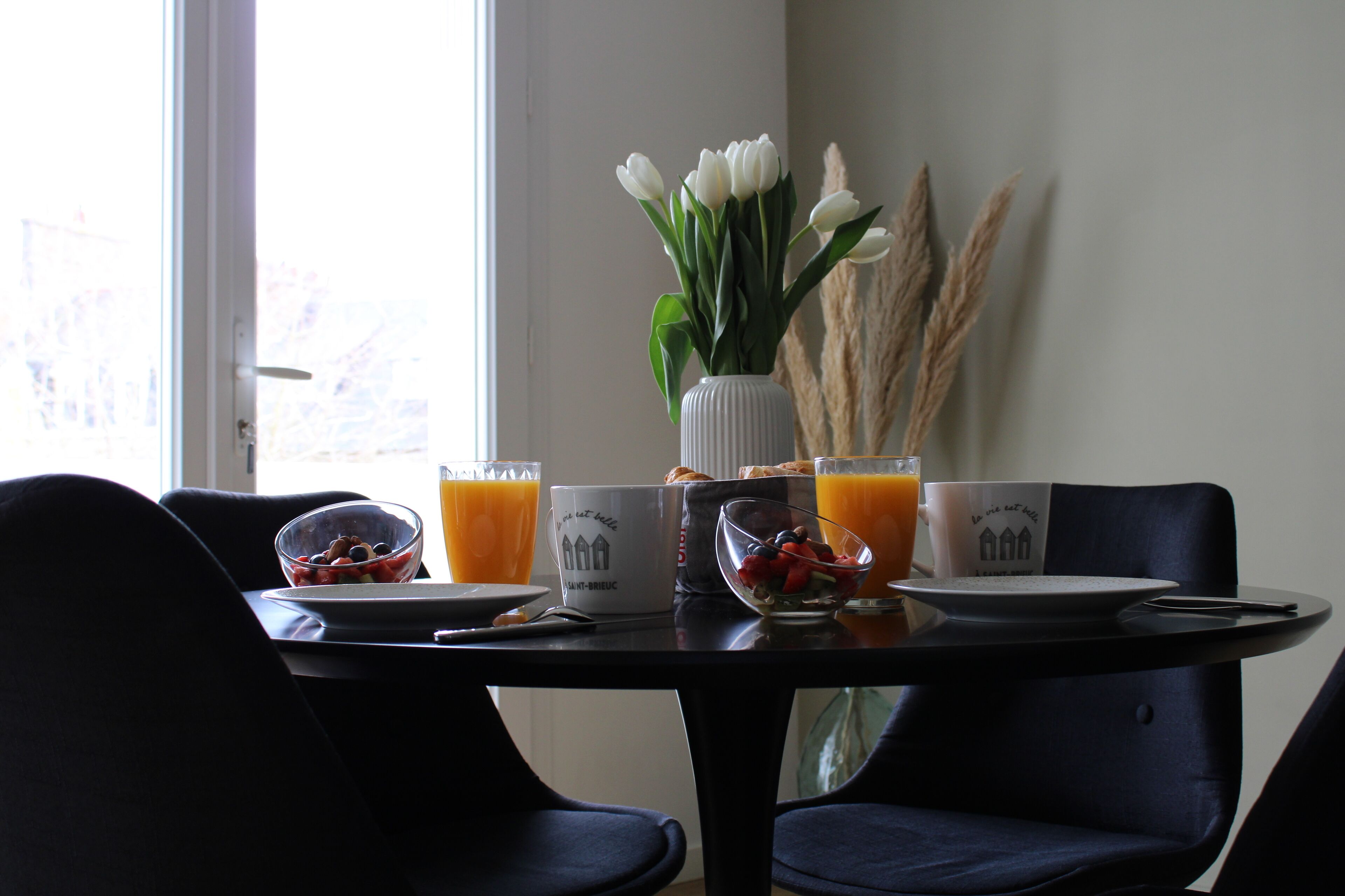 Mesa de jantar com itens de café da manhã, um vaso com flores brancas e uma janela com vista.