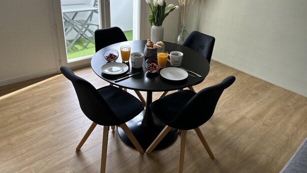 A dining table with four chairs, a vase with white flowers, and breakfast items including cereal, juice, and a croissant.