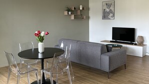A modern living room with a grey sofa, a round dining table with clear chairs, a black chandelier, and a flat-screen TV on a white stand.