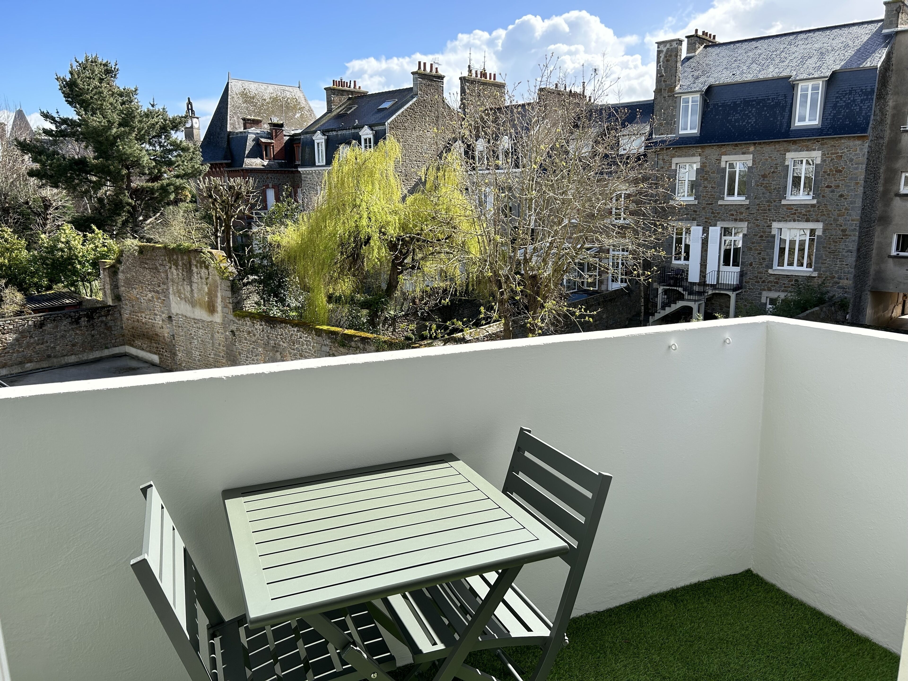 Un balcon avec une table et des chaises, donnant sur un quartier résidentiel avec des bâtiments en pierre et des arbres.