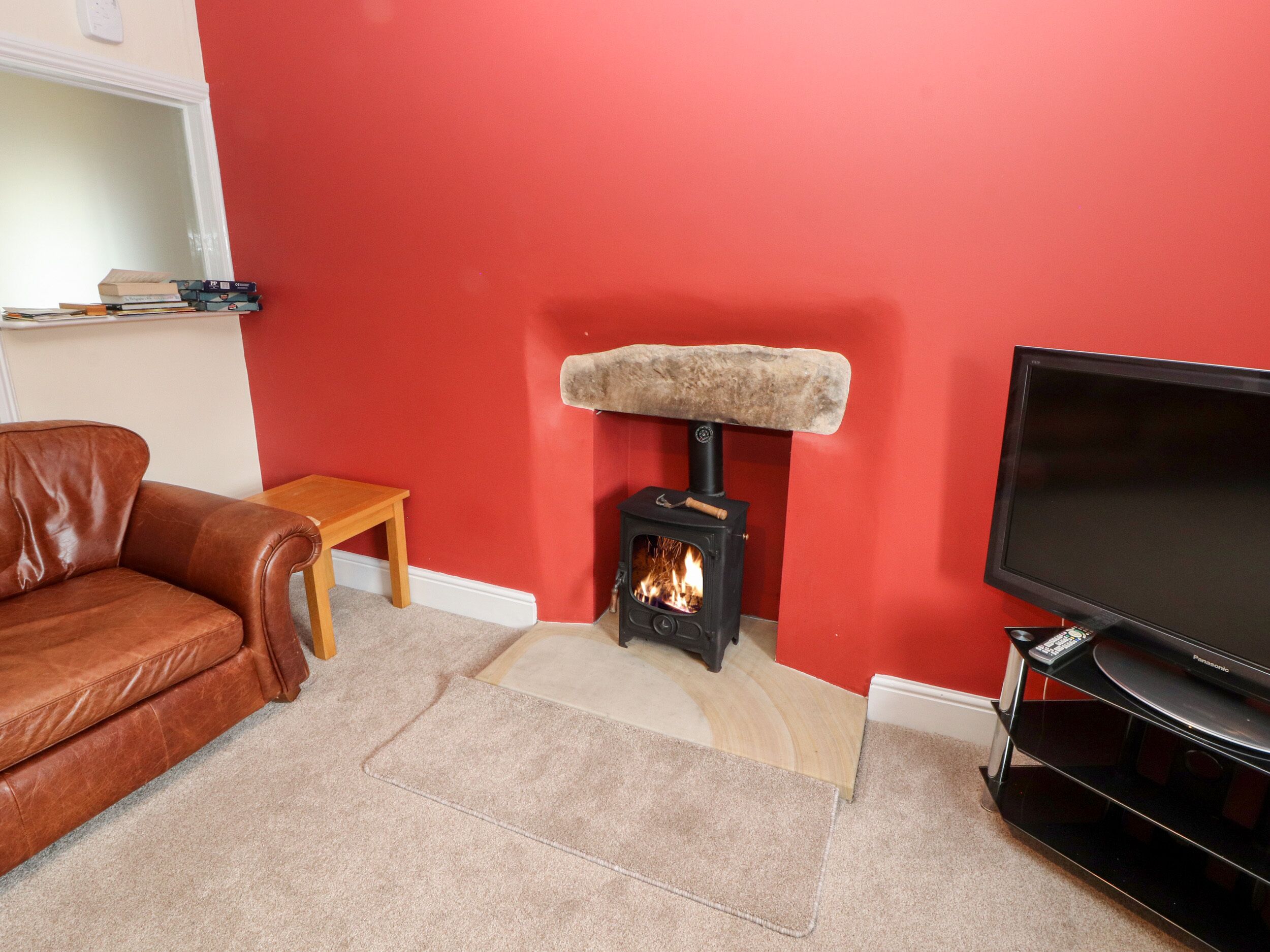A living room with a red wall, a brown leather sofa, a small wooden table, a stone cat bed, a black wood-burning stove, and a black television on a stand.