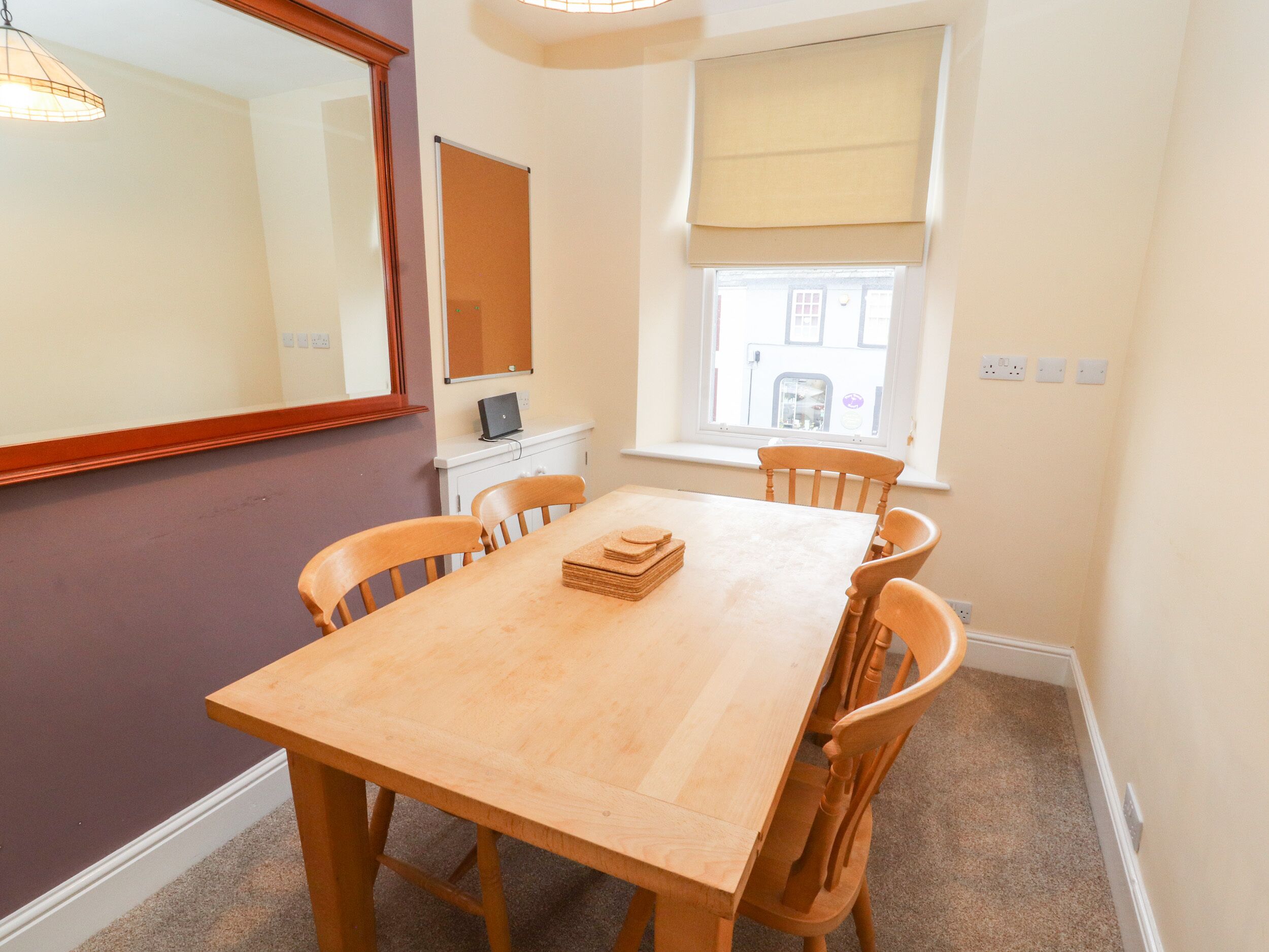 A dining area with a wooden table and chairs, a laptop on a small table, a mirror, and a window with a blind.