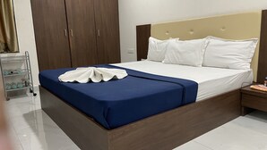 A neatly made bed with a blue bedspread and white pillows, a wooden wardrobe, and a small bedside table with a book and a bottle. - Hotel Millennium Plaza (Jamnagar)