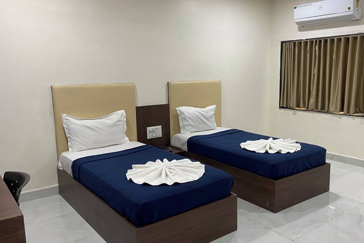 A hotel room with two beds, each with a headboard, a window with curtains, and an air conditioning unit on the wall.