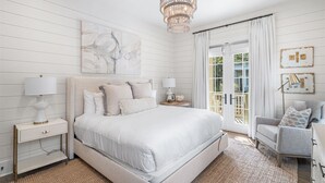 Une chambre à coucher avec un grand lit, des tables de chevet avec des lampes, un lustre, un tableau et une vue à travers une porte vitrée.