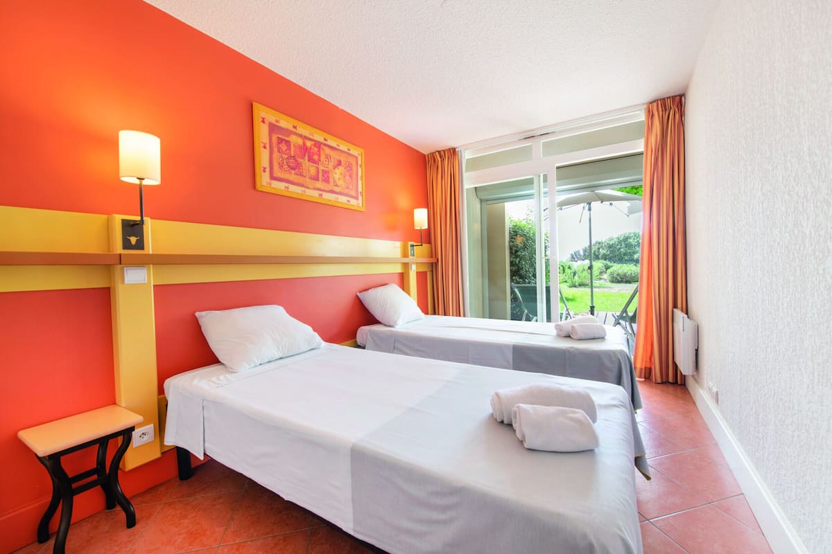 A hotel room with two beds, a small table, a lamp, and a view of the outdoors.