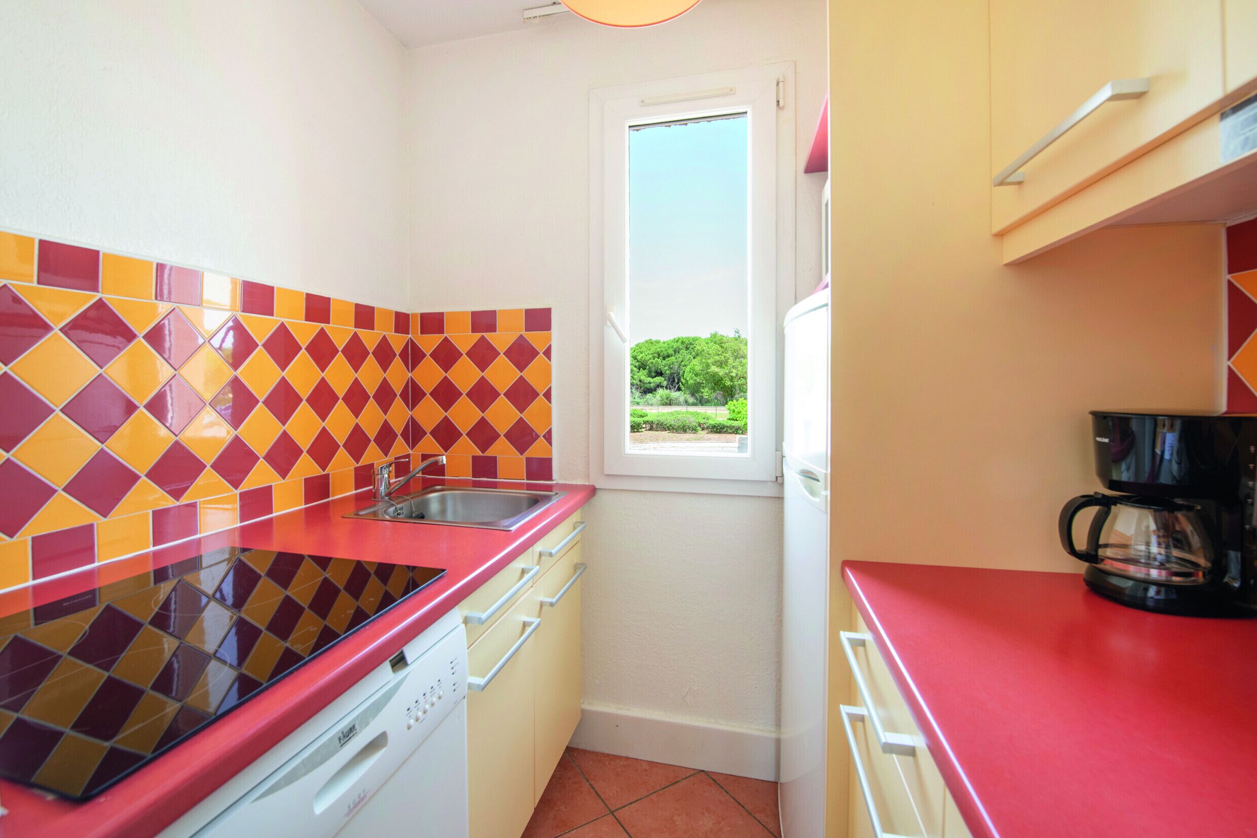 A compact kitchen with a red countertop, a window with a view of greenery, and a coffee maker.