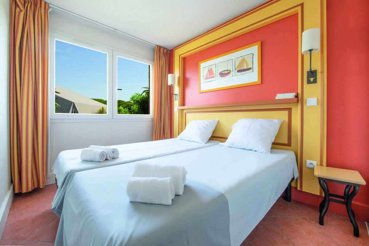 A hotel room with two beds, a window with curtains, a painting on the wall, and a small table.