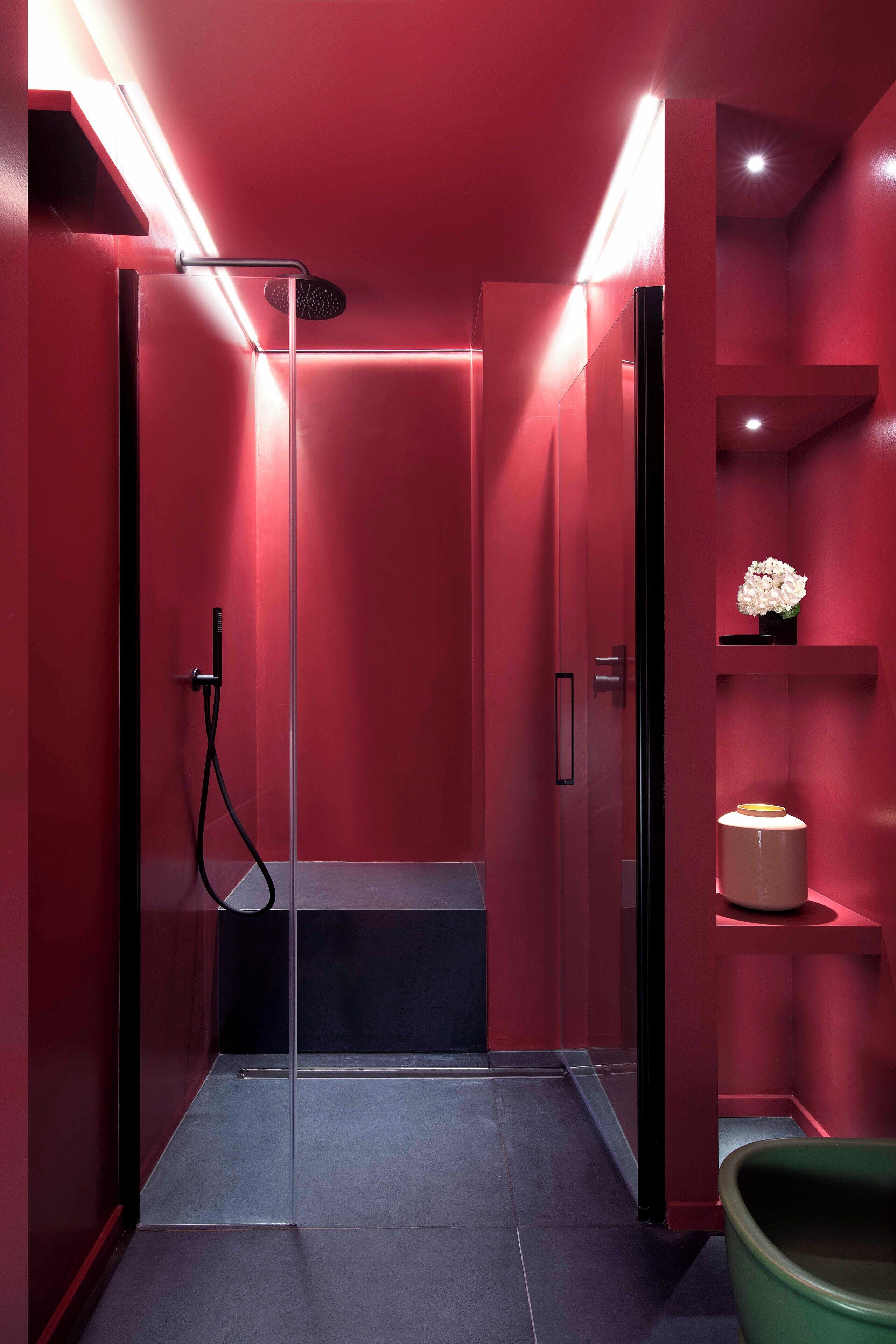 A modern bathroom with a glass-enclosed shower, red walls, and a green chair.