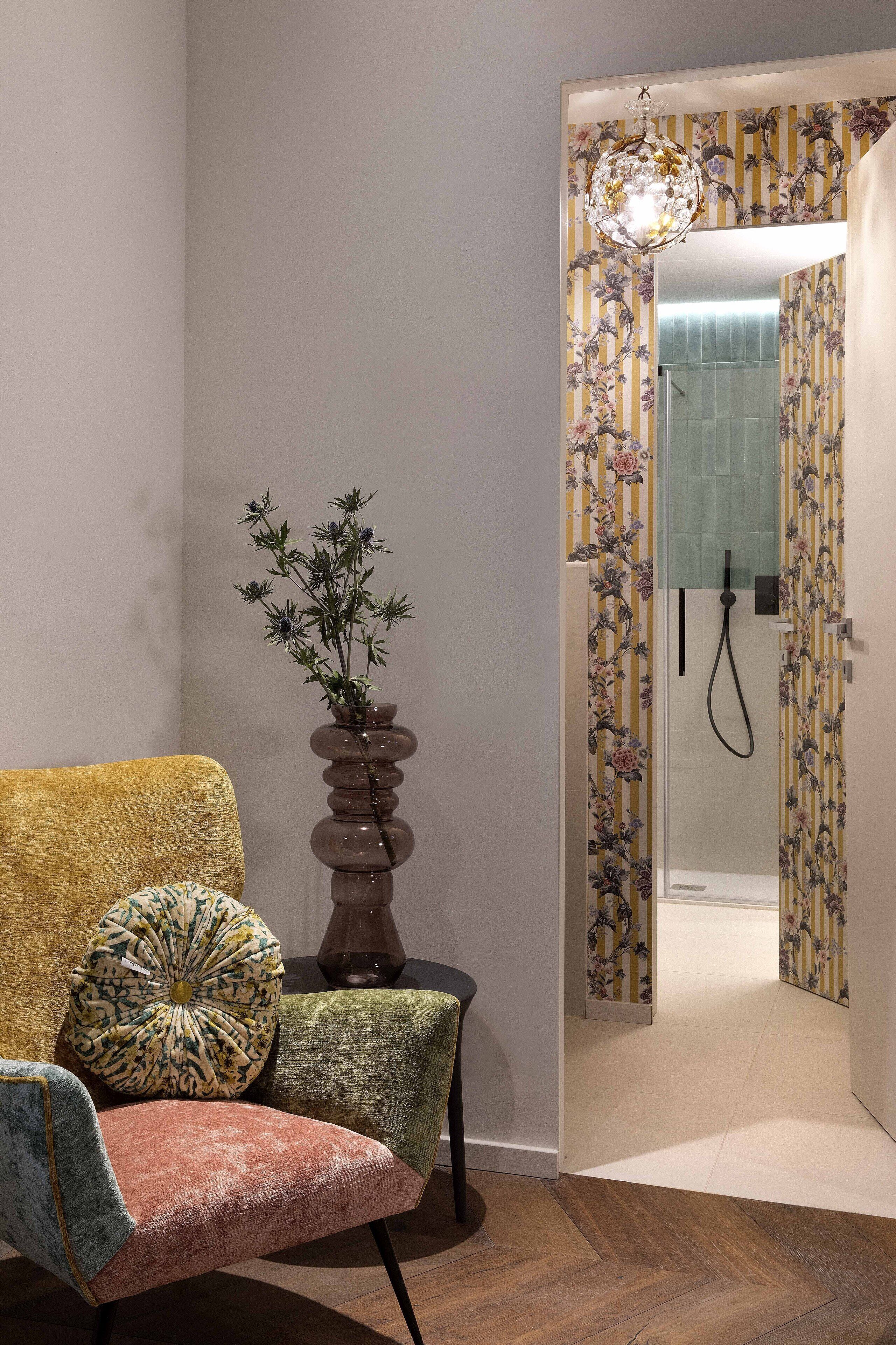 Una habitación con piso de madera, una silla con cojín estampado, un florero con flores secas y una zona de ducha con papel tapiz floral.
