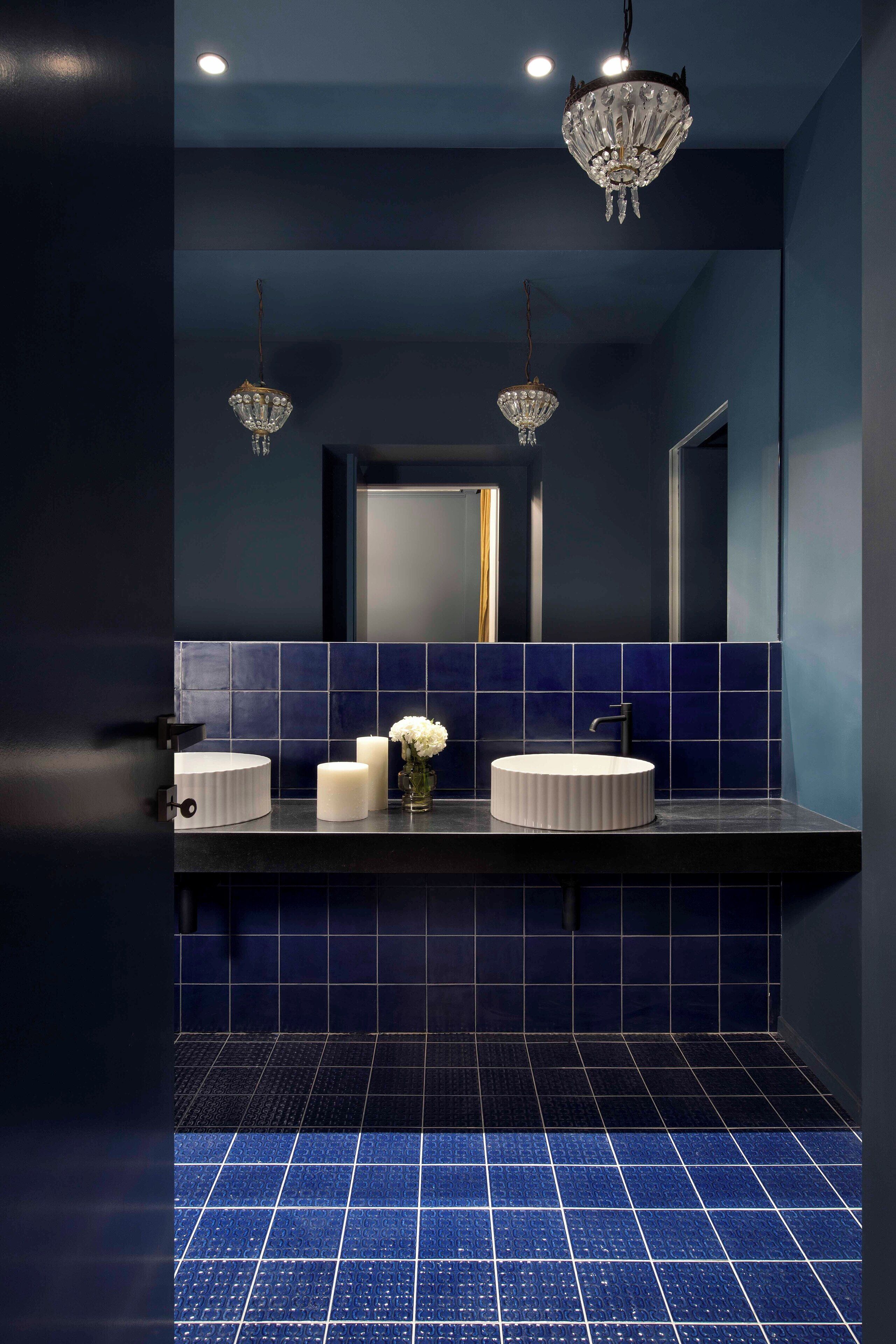 A modern bathroom with a dark blue tiled wall, a round white sink, and two crystal chandeliers.