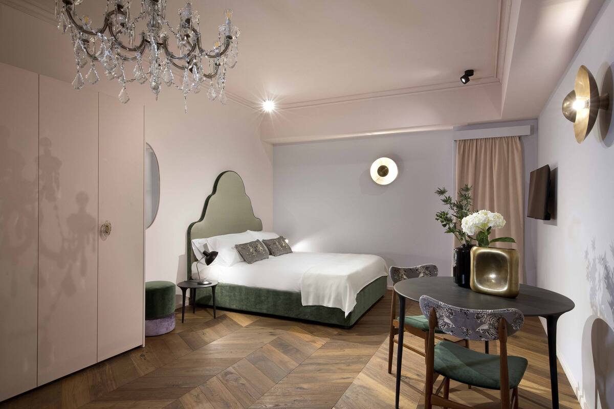 A bedroom with a bed, a chandelier, a round wall clock, a table with chairs, and a vase with flowers.