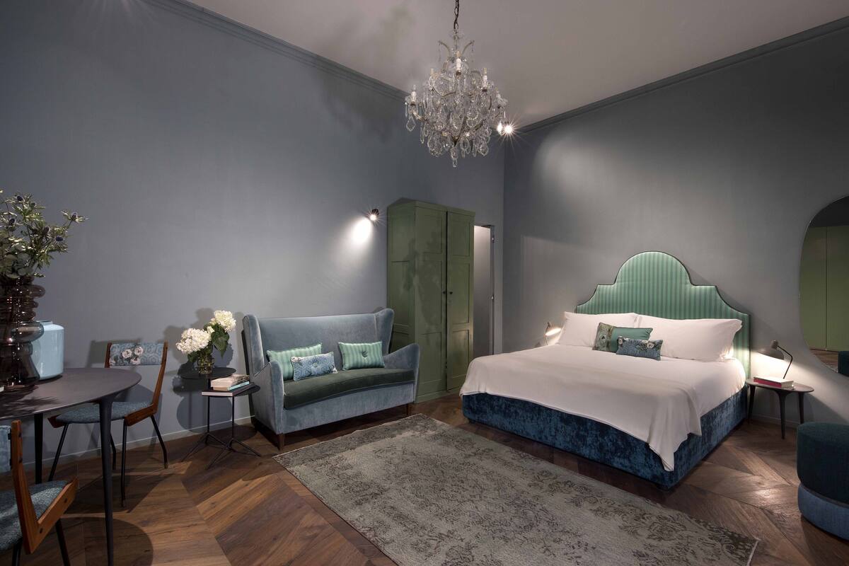 A bedroom with a bed, a green wardrobe, a blue sofa, a dining table with chairs, and a chandelier.