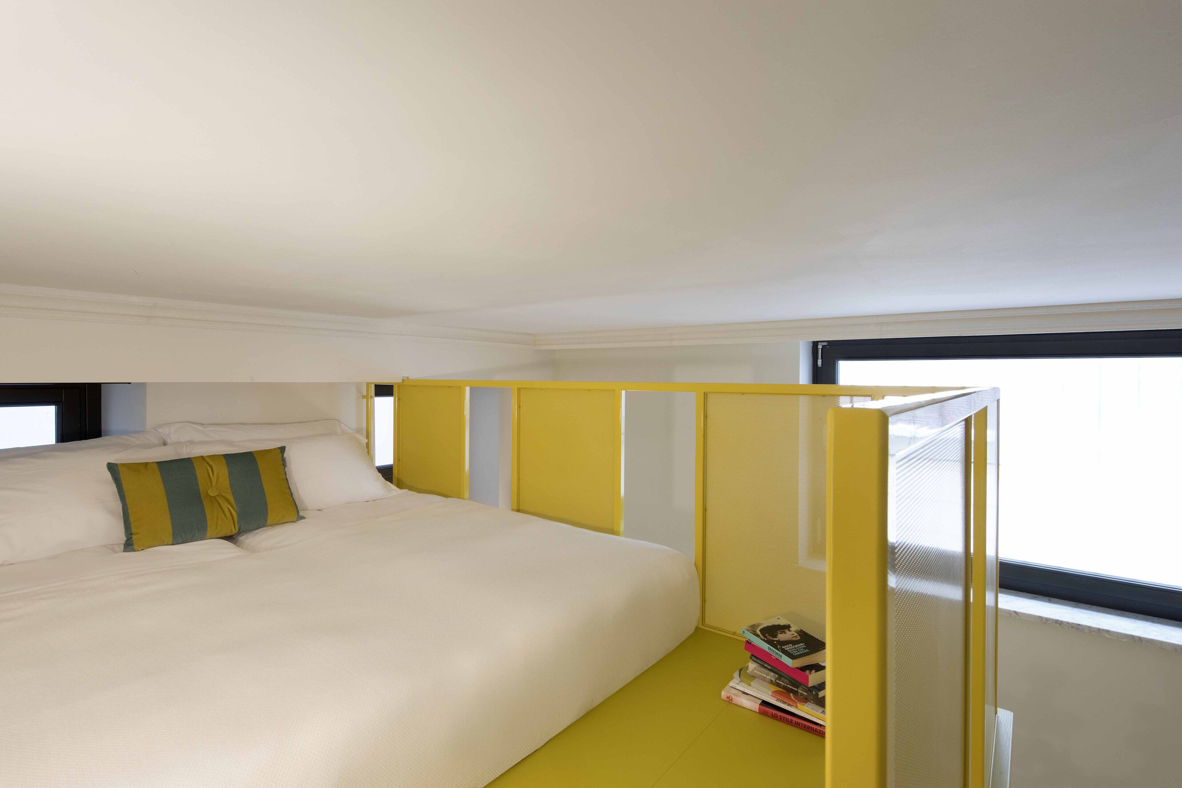 A bed with white bedding and a yellow headboard, a window with a view, and a stack of books on the floor.