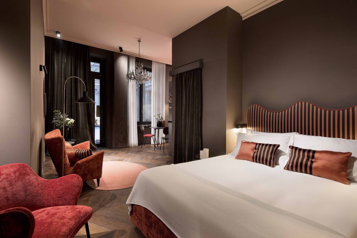 A hotel room with a large bed, red armchairs, a chandelier, and a view of the outside.