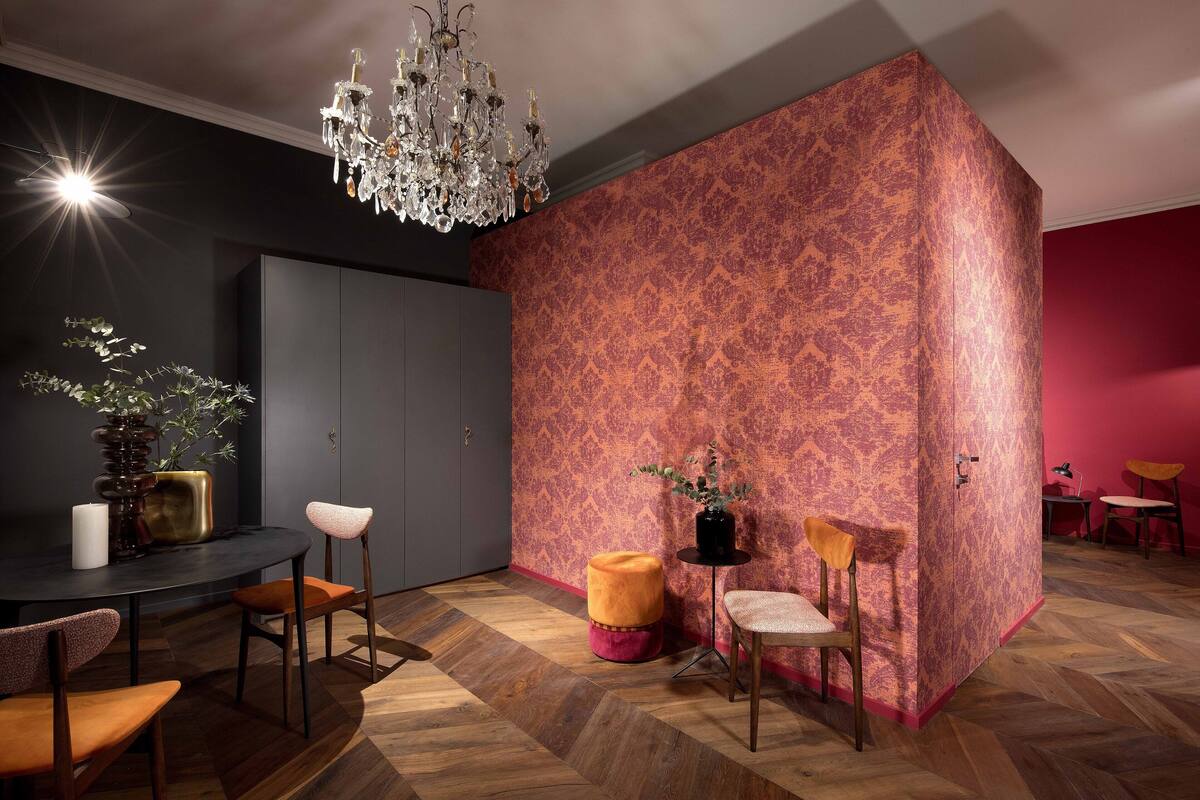 A room with a dark wall, a patterned accent wall, a chandelier, a round ottoman, and a dining area with chairs and a table.
