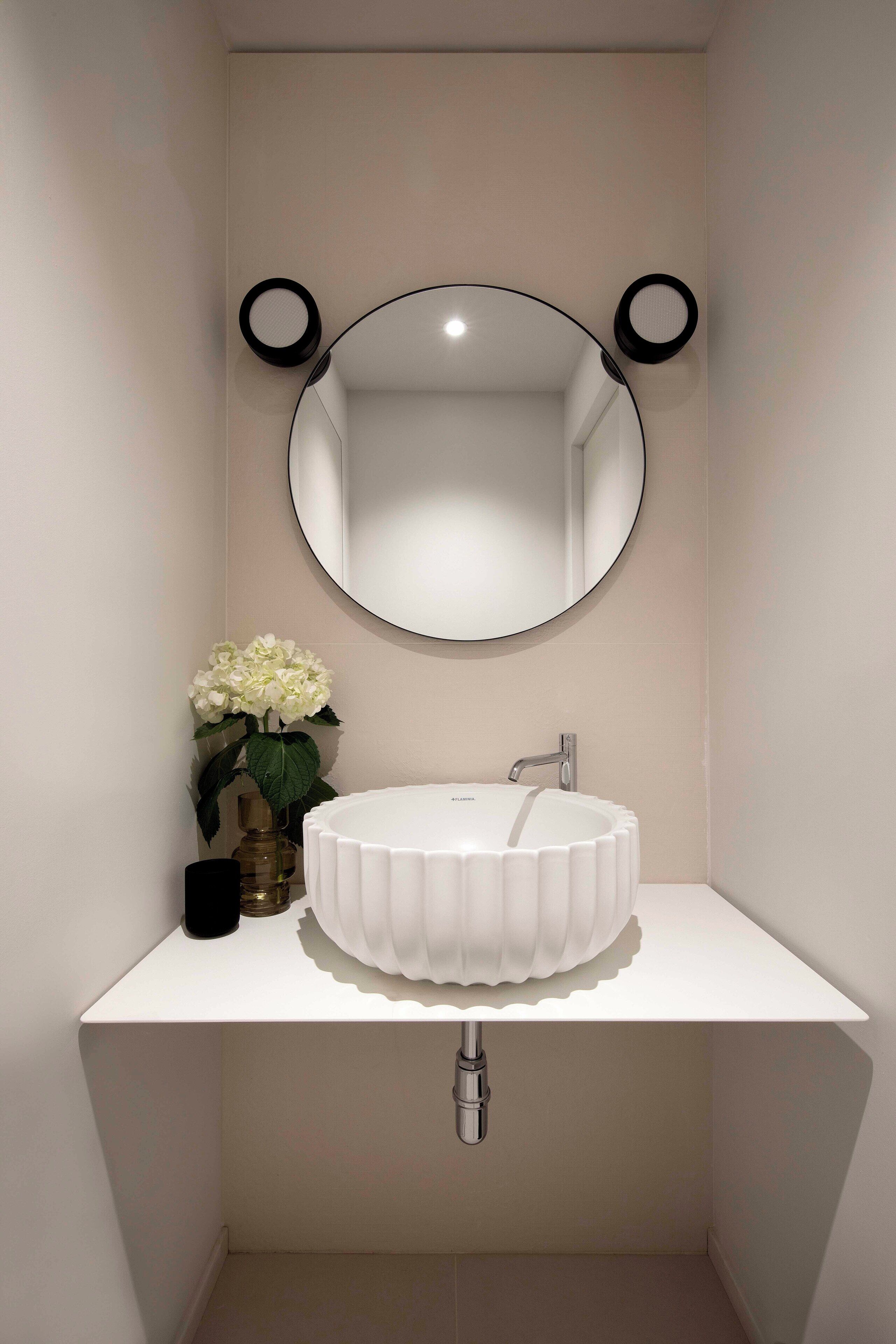 Ein Badezimmer mit einem weißen Waschbecken, einem runden Spiegel und einer Vase mit weißen Blumen.