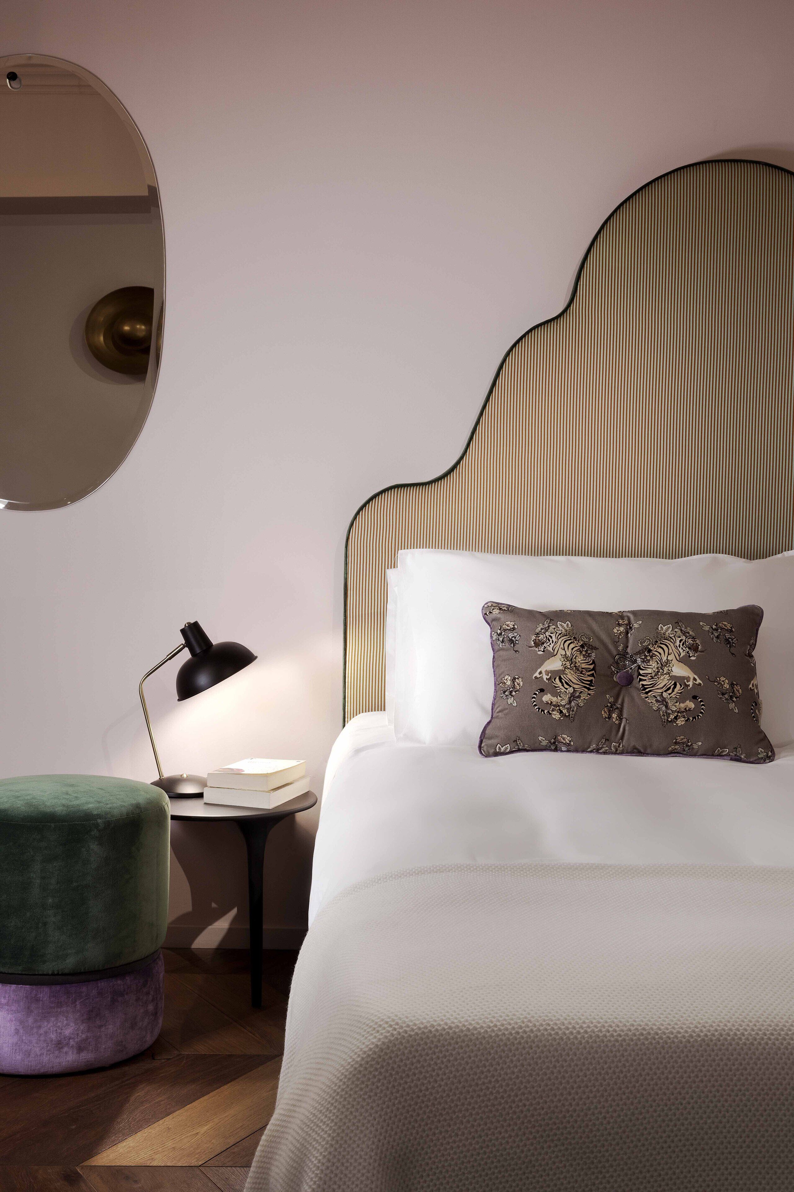 A neatly arranged bedroom with a large bed, a round mirror, a small side table, and a green ottoman.