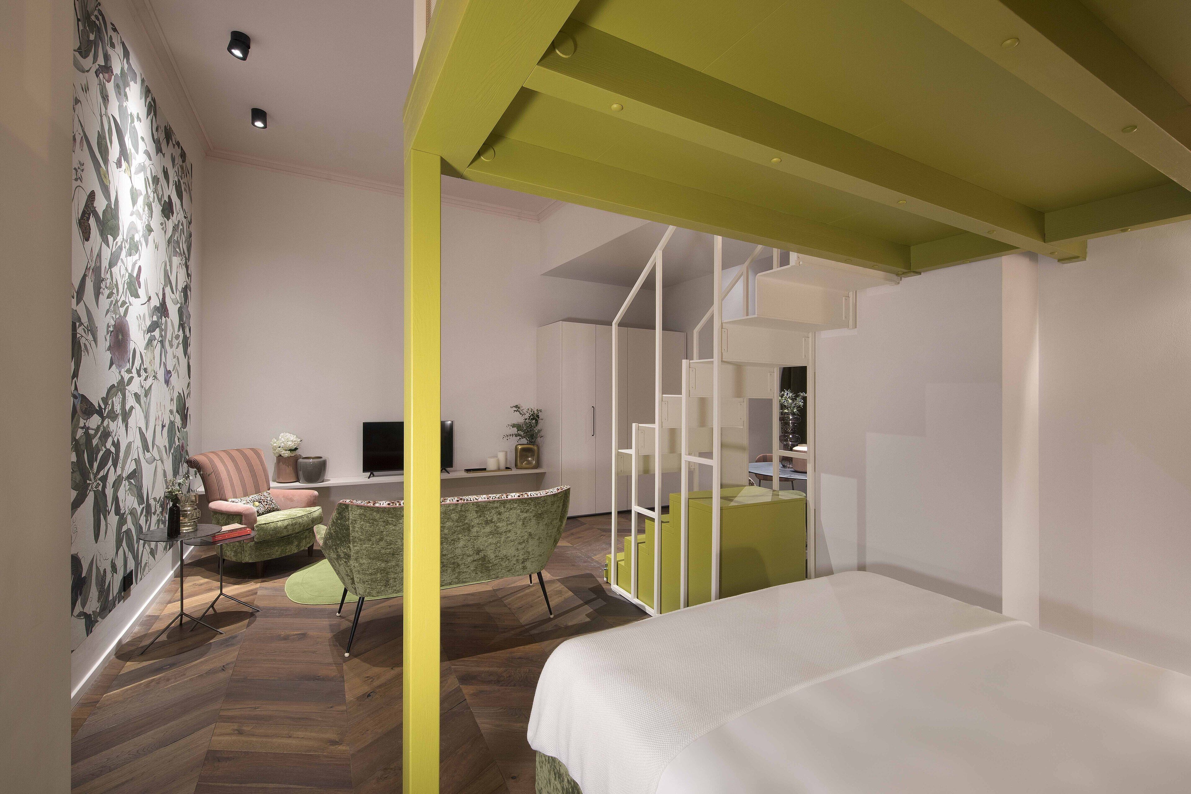 A modern interior with a bed, a green armchair, a small table, and a staircase leading to an upper level.