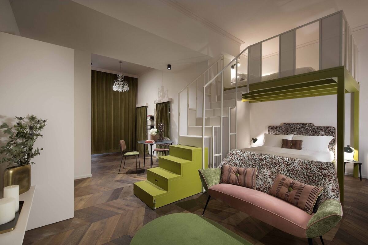 A modern hotel room with a bunk bed, a green staircase, a dining area, and a chandelier.
