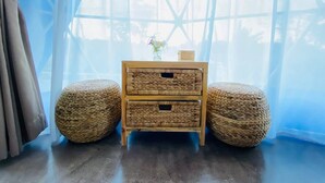 Quarto com mesa de cabeceira de madeira, dois pufes trançados, um vaso com flores e cortinas leves.