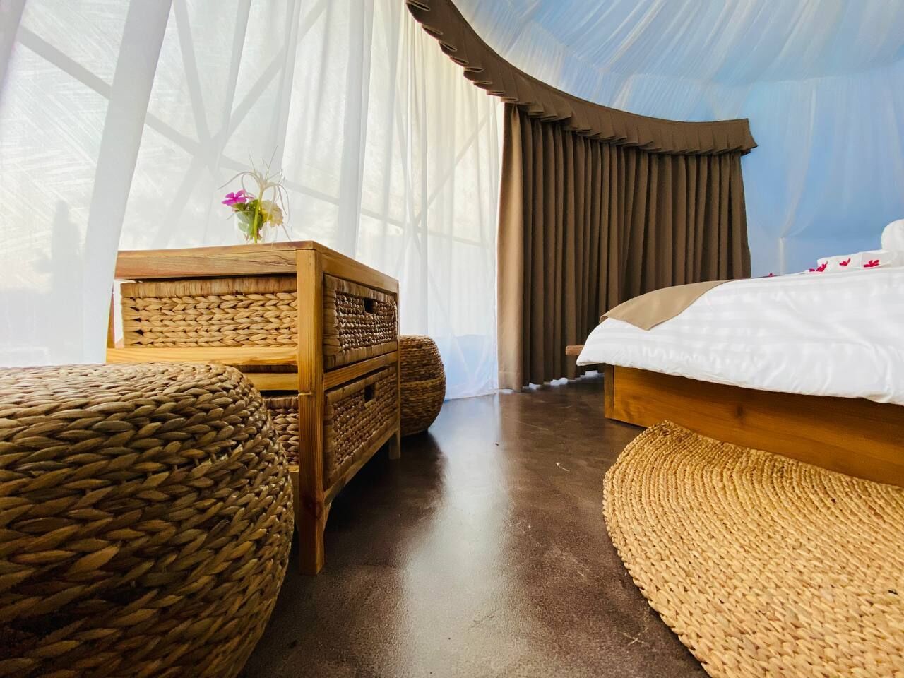 Una habitación con una cama, un aparador de madera y otomanas tejidas.