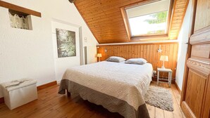 Un dormitorio con techo de madera, una cama grande con colcha estampada, dos lámparas de noche y vistas a través de un tragaluz.