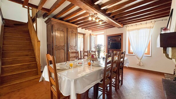 Comedor con mesa de madera dispuesta para cuatro personas, rodeada de sillas. Se ve una escalera a la izquierda y un televisor montado en la pared a la derecha.