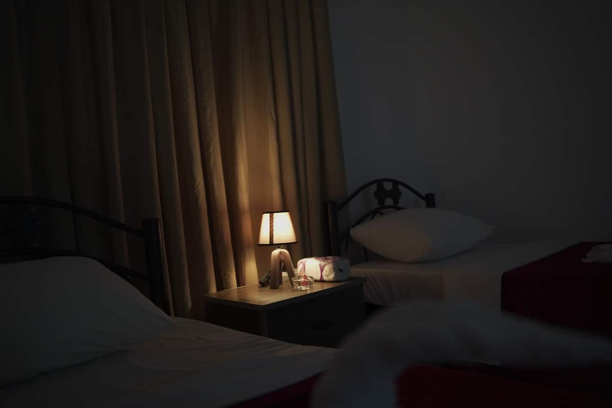 A dimly lit hotel room with a bed, a nightstand with a lamp, and a window with curtains.