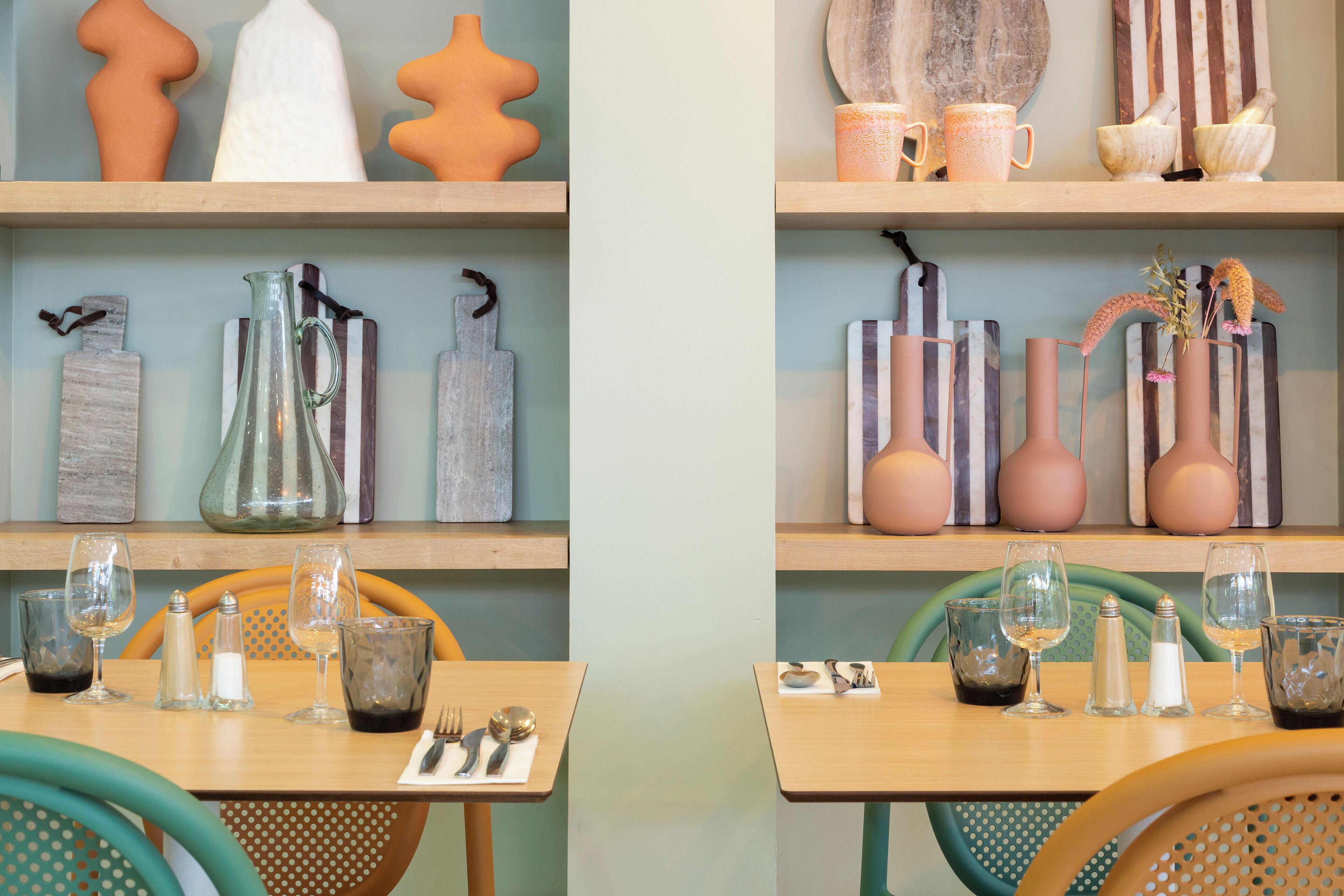 A dining area with wooden tables and chairs, glassware, and decorative items on shelves.