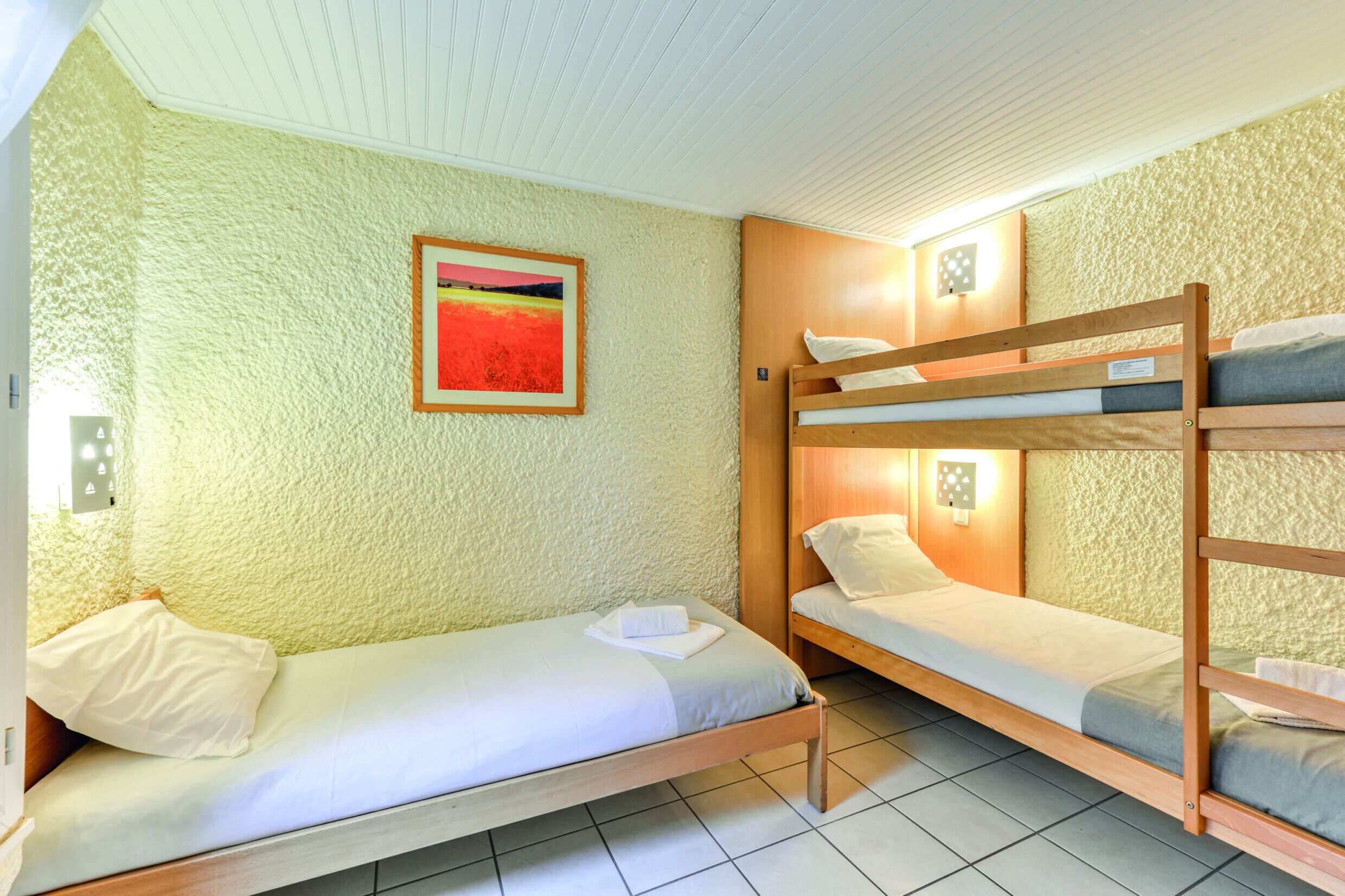 Een slaapzaal met stapelbedden, een enkel bed, een ingelijste foto aan de muur en een lichtschakelaar.