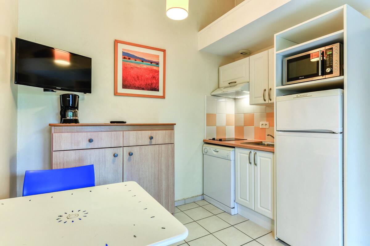 A compact kitchen with a microwave, dishwasher, and a coffee maker. There is a table with a blue chair, and a TV mounted on the wall.