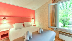 A hotel room with a large bed, a nightstand, a window with curtains, and a view of greenery outside. - SOWELL RESIDENCES Les Lauriers Roses (Agde)