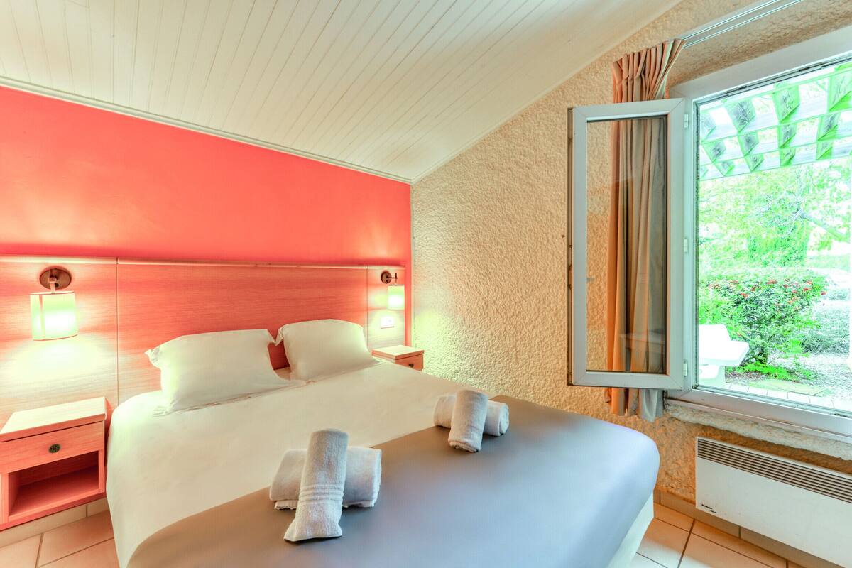 A hotel room with a large bed, a nightstand, a window with curtains, and a view of greenery outside.