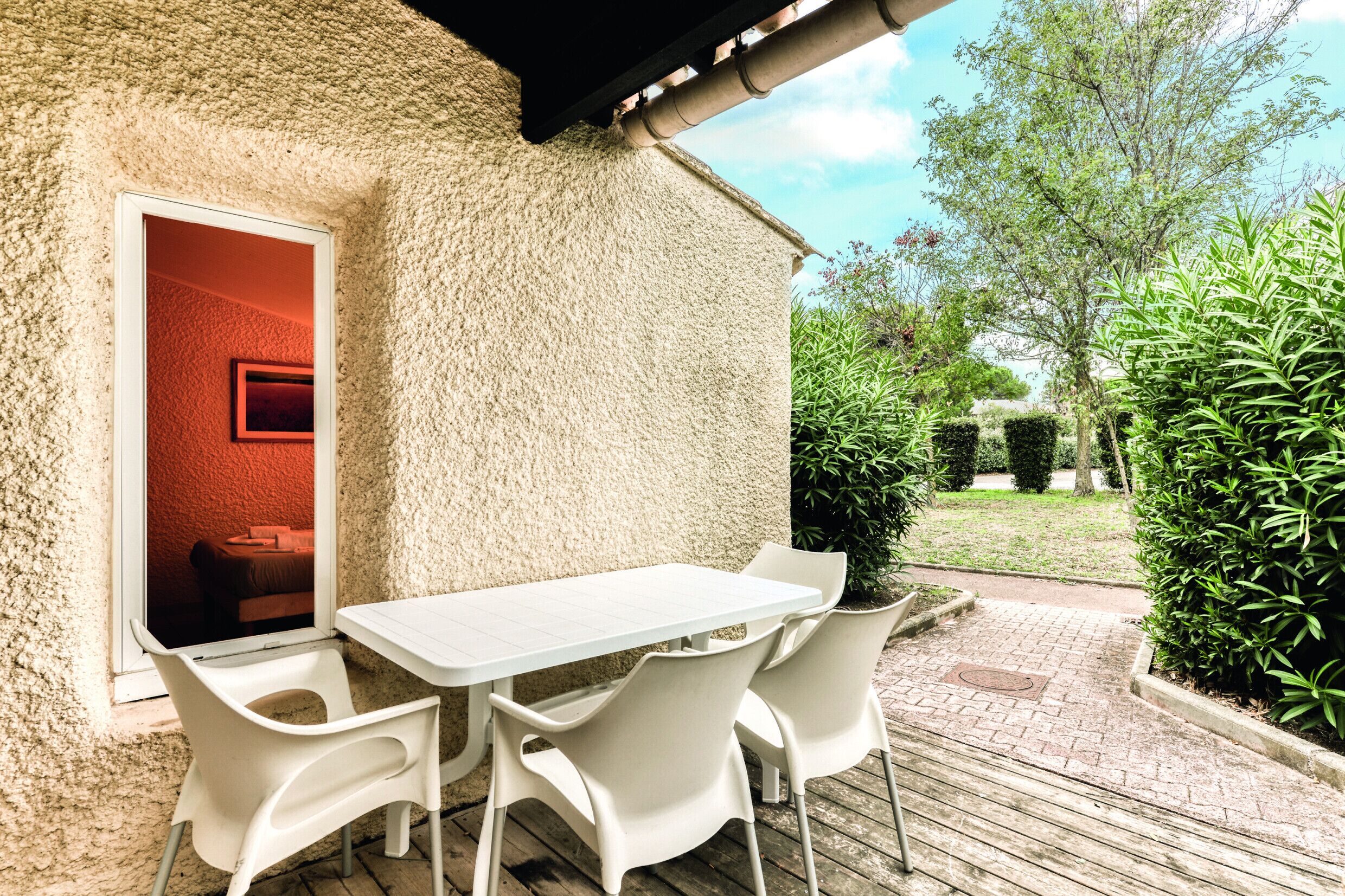 A patio with a white table and chairs, a red-framed window, and a view of a garden with hedges and trees.
