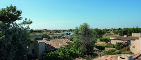 A residential area with terracotta-tiled roofs, a clear blue sky, and a distant view of a body of water.