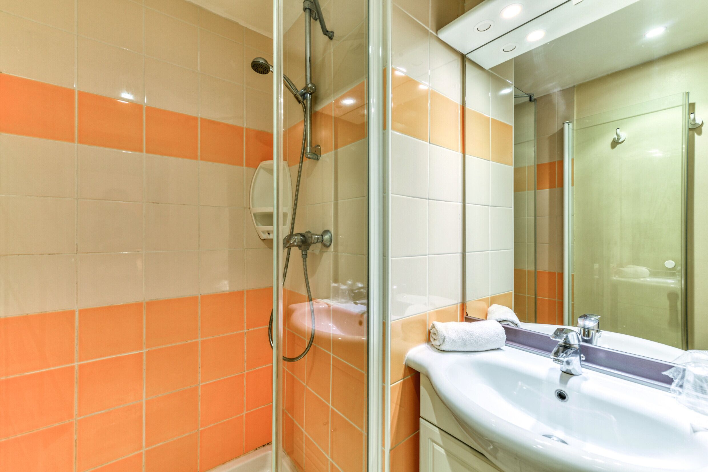 Une salle de bain avec une douche à parois de verre, un lavabo blanc et des murs recouverts de carreaux orange et blancs.