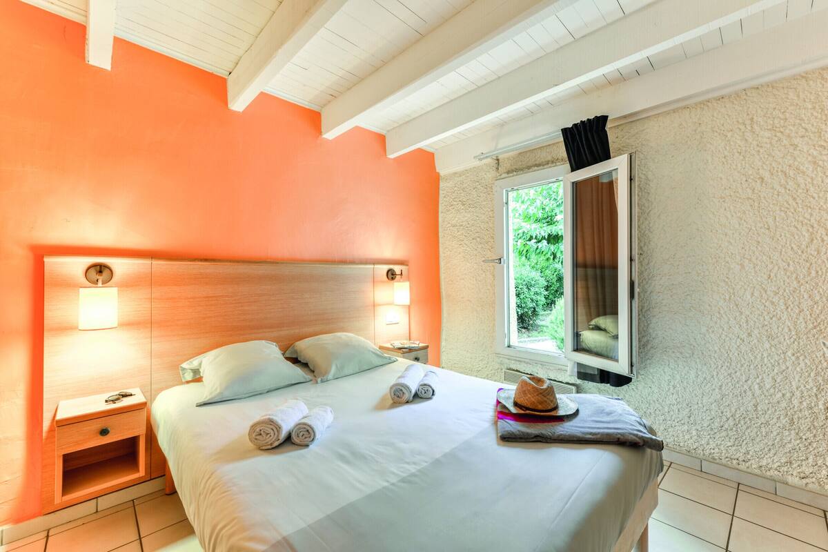 A hotel room with a large bed, two bedside tables, a window with a view of greenery, and a wooden ceiling.