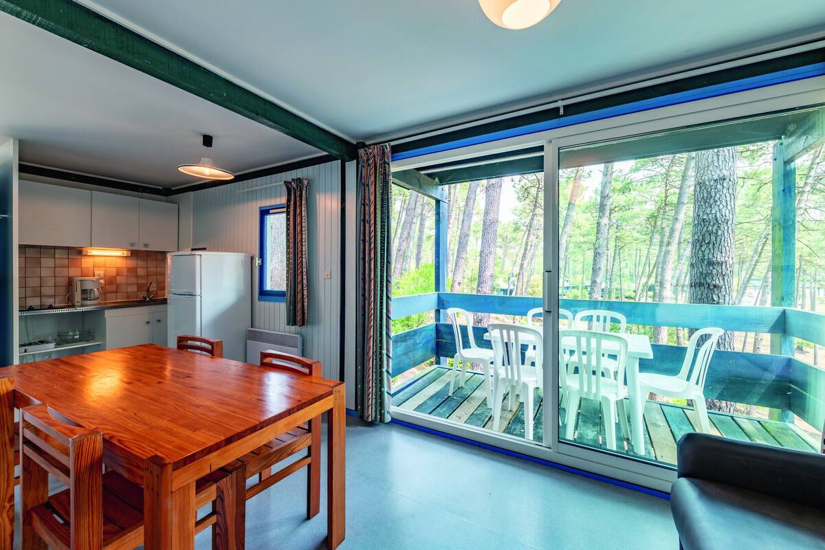 A modern kitchen with a wooden table and chairs, a refrigerator, and a dining area with a view of a balcony and trees.