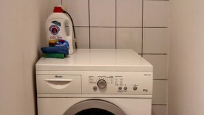 Une machine à laver à chargement frontal se trouve dans une buanderie, avec une bouteille de lessive et un chiffon posés sur le dessus.