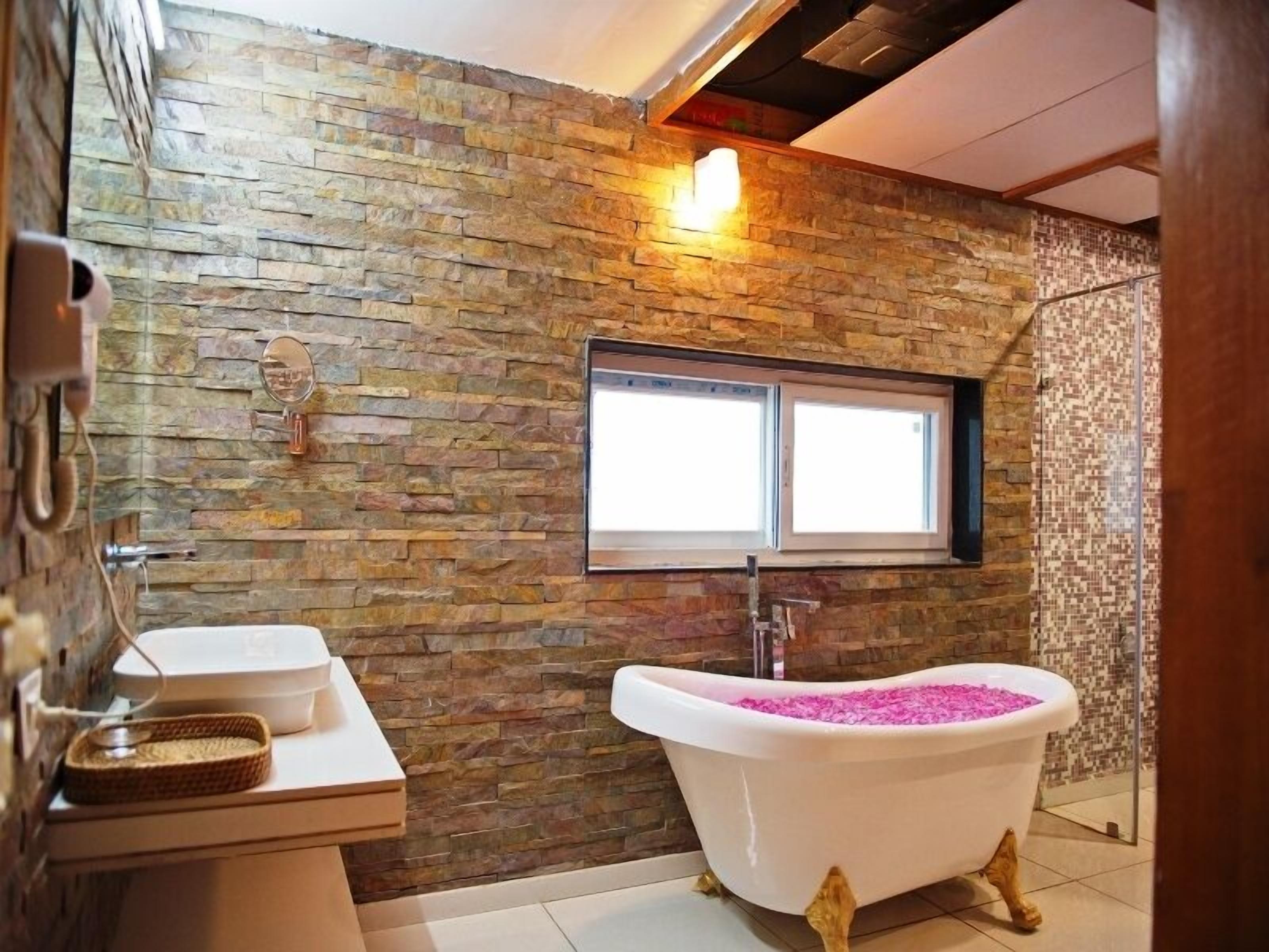 Un bagno con parete in pietra, una finestra, una vasca da bagno con sali da bagno rosa e un lavandino con specchio.