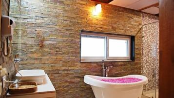 Un bagno con parete in pietra, una finestra, una vasca da bagno con sali da bagno rosa e un lavandino con specchio.