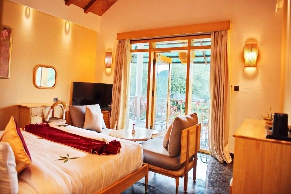 Habitación de hotel con cama, escritorio, silla, televisor y balcón con cortinas.