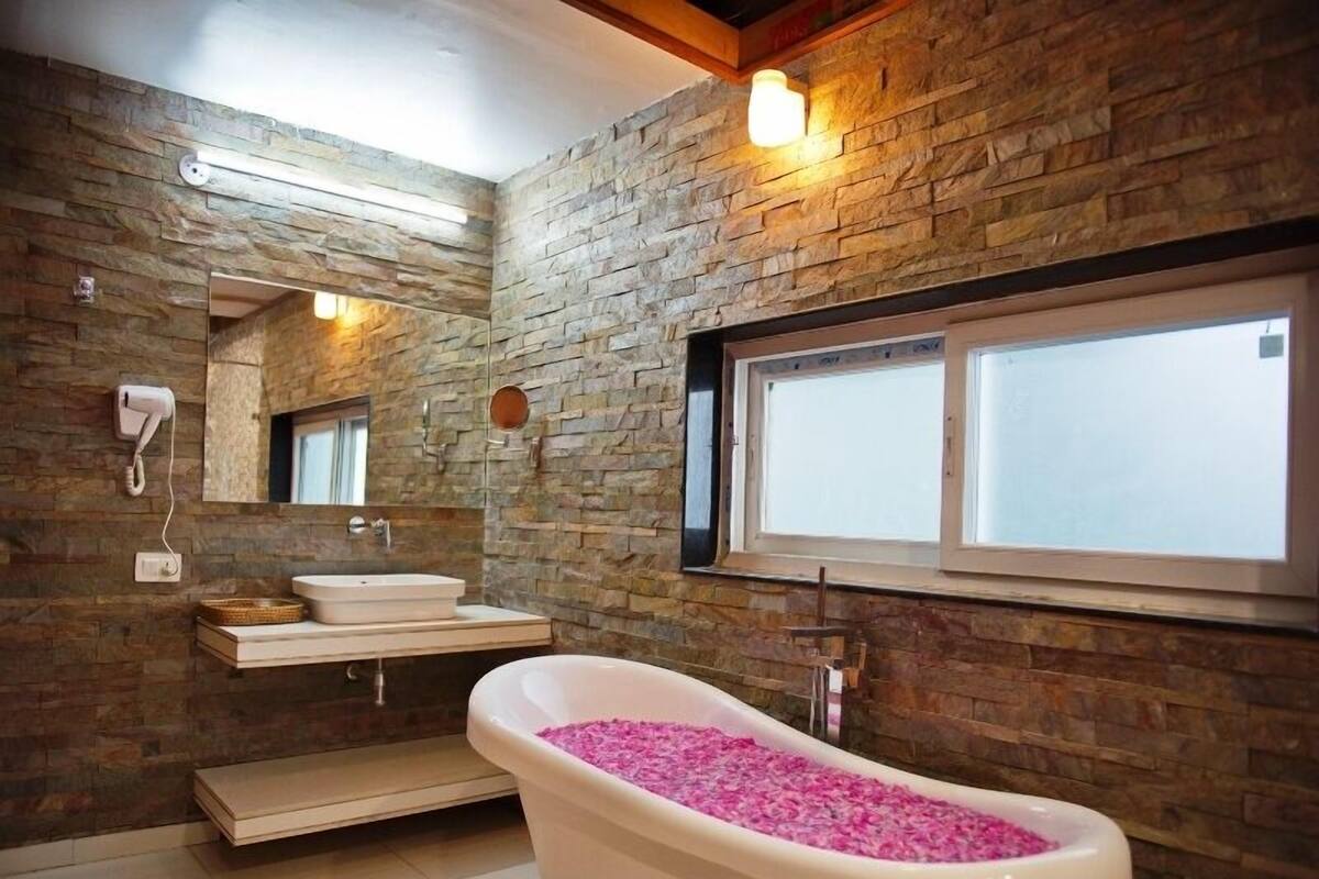 Un baño con pared de piedra, una bañera independiente llena de productos de baño rosados, un lavabo y una ventana sobre el lavabo.