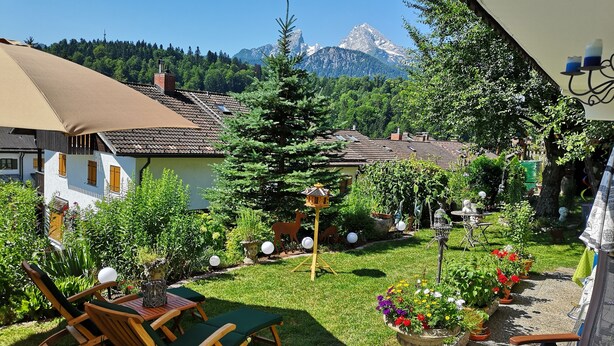 Un patio con mobiliario de madera, un jardín con plantas en macetas y una vista a la montaña al fondo.