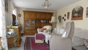 Una sala de estar con un sofá color beige, un tapete estampado, un mueble de madera con puertas de cristal y una araña.