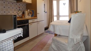 Cocina compacta con estufa, horno y microondas. Hay una cama con cubre cama blanco y una silla. La habitación tiene una ventana con cortinas.